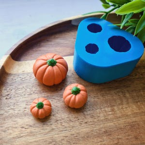Op de afbeelding: Een felblauwe siliconen mal met drie pompoenvormige holtes naast drie kleine, oranje, realistische pompoenfiguurtjes met groene steeltjes, die het eindresultaat tonen dat met de verschillende maten van de mal is gemaakt.