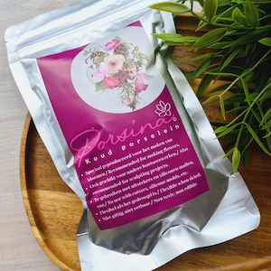 Puede incluir: Una bolsa plateada con una etiqueta rosa que dice "Porsina Koud Porselein". La etiqueta tiene un diseño floral y un texto que describe el producto como porcelana fría para hacer flores y otras manualidades.