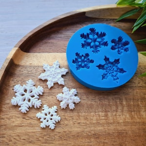 May include: A blue silicone mold with six snowflake shapes. Five white snowflake-shaped decorations are on a wooden surface next to the mold.