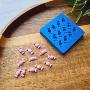 May include: A square blue silicone mold featuring twelve recessed candy-shaped cavities, accompanied by twelve small, light pink, three-dimensional candy-shaped pieces scattered nearby, each with a distinct wrapped-style design.