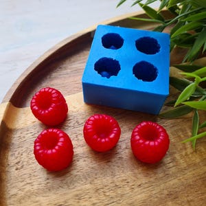 Puede incluir: Cuatro caramelos rojos en forma de frambuesa sobre una superficie de madera junto a un molde de silicona azul con cuatro cavidades redondas.