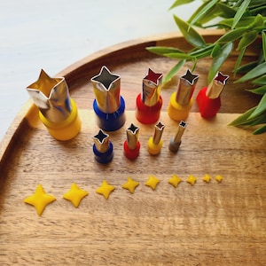 Pode incluir: Um conjunto de nove cortadores de argila metálicos em forma de estrela com cabos de plástico coloridos em vermelho, azul e amarelo. Os cortadores variam em tamanho e estão dispostos em duas filas. Abaixo, nove estrelas amarelas recortadas.