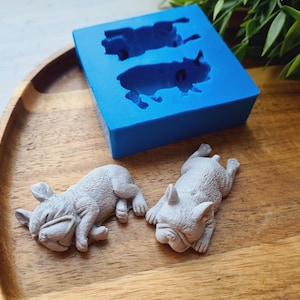 May include: A bright blue silicone mold featuring two recessed cavities shaped like sleeping French bulldogs, accompanied by two light gray, detailed figurines of sleeping French bulldogs that have been cast from the mold.