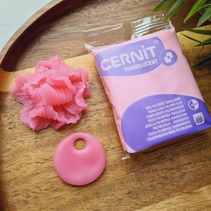 May include: A wooden tray holds a pink polymer clay flower, a pink pendant with a hole, and a package of translucent polymer clay. The package is labeled "CERNIT TRANSLUCENT". Green leaves are in the background.