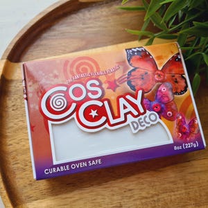 Può includere: Una scatola rettangolare di Cosclay Deco, un materiale modellabile in plastica flessibile. La confezione presenta sfumature arancioni e viola, illustrazioni di farfalle e il testo: The Fantastic Flexible Plastic, Cosclay Deco, Curable Oven Safe, 8oz (227g).