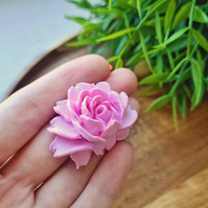 May include: A pink, handmade, polymer clay rose flower. The rose is detailed with multiple layers of petals.