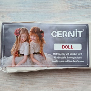 May include: A package of white modeling clay with a porcelain finish. The package features a photo of two girls wearing white dresses and floral headbands. The text on the package reads "CERNIT DOLL Modelling clay with porcelain finish Pâte à modeler finition porcelaine Modelliermasse mit Porzellanschimmer."