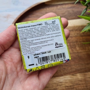 May include: A yellow and black package of FIMO modelling clay. The package has a barcode and the text "Veido&scaron;anas masa FIMO" in Latvian. The text "schwarz/black/noir" and "negro" are also visible on the package.