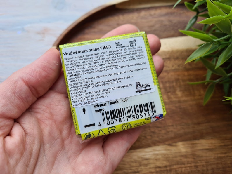 May include: A yellow and black package of FIMO modelling clay. The package has a barcode and the text "Veido&scaron;anas masa FIMO" in Latvian. The text "schwarz/black/noir" and "negro" are also visible on the package.
