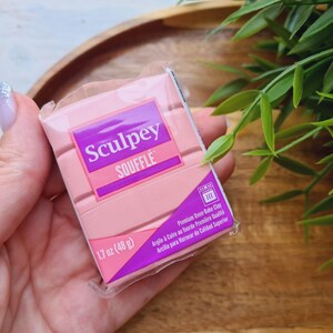 May include: A package of pink Sculpey Souffl&eacute; polymer clay, 1.7 oz (48 g), held in a hand. The packaging features the brand name in white and purple, with text indicating it's premium oven-bake clay.