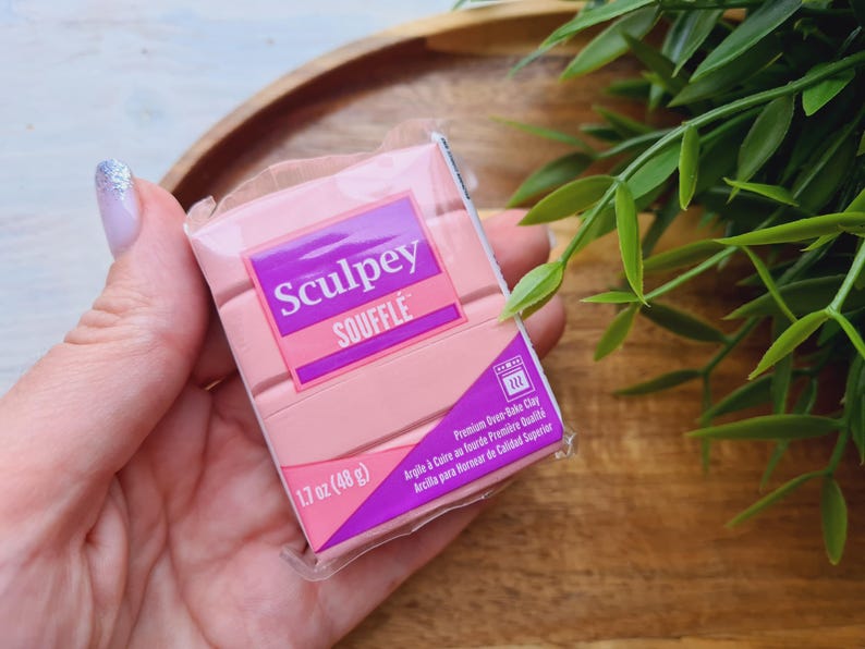 May include: A package of pink Sculpey Souffl&eacute; polymer clay, 1.7 oz (48 g), held in a hand. The packaging features the brand name in white and purple, with text indicating it's premium oven-bake clay.