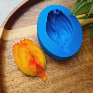 May include: A bright yellow and orange tulip flower next to a blue silicone mold in the shape of a tulip. The items are on a wooden tray with a green plant in the background.