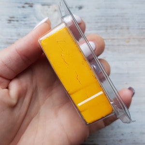 Può includere: Un blocco rettangolare di materiale giallo brillante, forse cera o argilla, racchiuso in una confezione di plastica trasparente. Il materiale ha una superficie leggermente strutturata e una striscia bianca. La confezione è tenuta in mano.