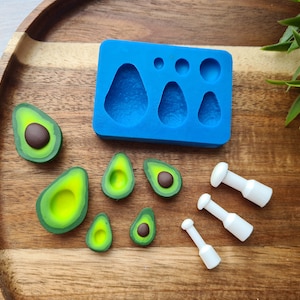 Puede incluir: Un molde de silicona azul con cuatro formas de aguacate y tres círculos pequeños. Hay seis piezas de arcilla polimérica en forma de aguacate, algunas con centros marrones, y tres herramientas blancas sobre una superficie de madera.