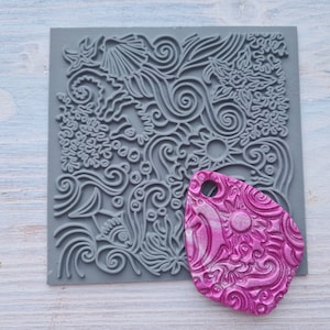 Puede incluir: Un sello de silicona gris con un diseño detallado de líneas en espiral, conchas marinas y otros elementos con temática oceánica. Un colgante de arcilla polimérica rosa con el mismo diseño se muestra junto al sello.
