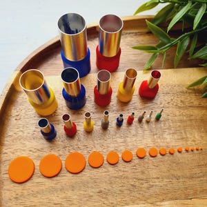Può includere: Una collezione di utensili perforatori in metallo con basi in plastica colorata di varie dimensioni, disposti in file. Il set include punzoni cilindrici più grandi con basi blu, rosse e gialle, insieme a punte metalliche più piccole e una fila di dischi circolari arancioni.