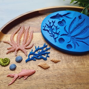 Pode incluir: Um molde de silicone azul redondo com desenhos em relevo de peixes, corais e conchas, acompanhado por várias figuras moldadas em rosa, azul, verde e bronzeado, incluindo um peixe, ramos de coral e várias conchas pequenas.