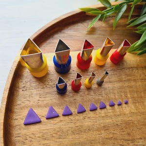 Puede incluir: Un juego de nueve cortadores de arcilla metálicos con forma de triángulo y bases de plástico de colores amarillo, azul, rojo y gris, dispuestos en dos filas, acompañados de nueve pequeños recortes de arcilla triangulares morados de varios tamaños.