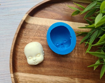 May include: A bright blue silicone mold shaped like a doll head and a matching off-white, smooth doll head casting. The mold features a recessed facial structure with defined eyes, nose, and mouth, while the casting shows the raised facial features.