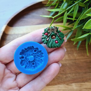 Puede incluir: Un molde de silicona azul para hacer una corona de Navidad. El molde tiene la forma de una corona con un lazo y bayas. El molde se muestra junto a una corona de Navidad verde y roja.