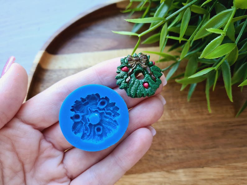 Puede incluir: Un molde de silicona azul para hacer una corona de Navidad. El molde tiene la forma de una corona con un lazo y bayas. El molde se muestra junto a una corona de Navidad verde y roja.