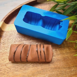 May include: A blue silicone mould for making chocolate bars. The mould is shaped like a chocolate bar with ridges. A chocolate bar with chocolate stripes is sitting next to the mould.