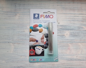 Puede incluir: Un paquete de 25 agujas para perforar cuentas para arcilla polimérica. El paquete es blanco con acentos azules y rojos y presenta el logotipo de FIMO. Las agujas son plateadas y vienen en un tubo de plástico transparente. El paquete también incluye instrucciones para usar las agujas.
