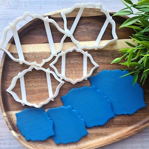 Könnte beinhalten: Weiße Ausstecher aus Kunststoff in dekorativer Form, zusammen mit blauen Fondant-Ausstichen, auf einem Holztablett angeordnet. Grünblättrige Pflanze im Hintergrund. Ideal zum Backen und Basteln.