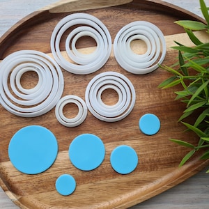 Puede incluir: Un juego de 8 cortadores de galletas redondos de plástico blanco de varios tamaños en una bandeja de madera con 5 círculos redondos azules.