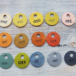 Puede incluir: Una colecci&oacute;n de 12 muestras de papel redondas en varios colores, incluyendo beige, amarillo, naranja, marr&oacute;n, verde, azul y gris. Cada muestra tiene un agujero perforado en el centro y un n&uacute;mero escrito en &eacute;l, como "050", "053" y "054".