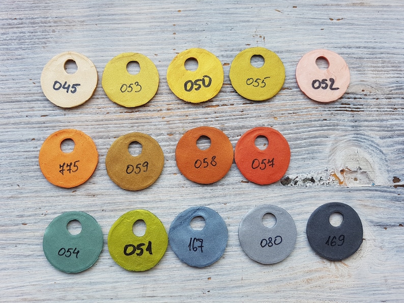 Puede incluir: Una colecci&oacute;n de 12 muestras de papel redondas en varios colores, incluyendo beige, amarillo, naranja, marr&oacute;n, verde, azul y gris. Cada muestra tiene un agujero perforado en el centro y un n&uacute;mero escrito en &eacute;l, como "050", "053" y "054".