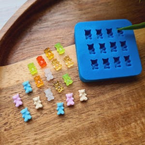 Peut inclure: Un moule en silicone bleu avec 16 cavités en forme d'ours, à côté d'une variété de bonbons gélifiés en forme d'ours de couleurs différentes. Les bonbons sont disposés en arc-en-ciel, avec certains bonbons de couleur bleu clair, rose et blanc.