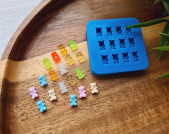 Könnte beinhalten: Eine blaue Silikonform mit 16 Bären-förmigen Vertiefungen, neben einer Vielzahl von bunten Gummibärchen. Die Gummibärchen sind in einem Regenbogenmuster angeordnet, wobei einige der Gummibärchen in hellblau, rosa und weiß gefärbt sind.