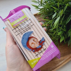 May include: A package of Sculpey Living Doll polymer clay in Light Claro | Clair color. The package is pink and yellow with a picture of a sleeping doll with a crescent moon. The package says "Semi-translucent, smooth blending, and perfect for fine detail." It also says "1 lb (454g)".