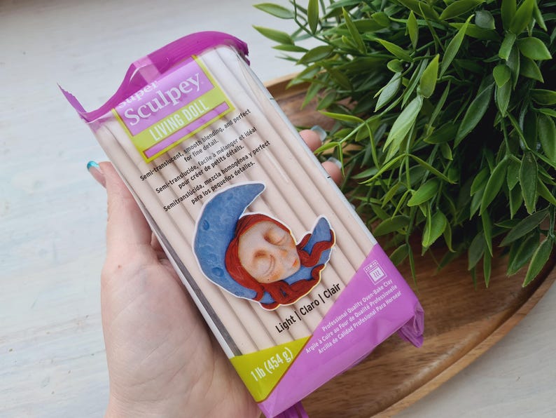 May include: A package of Sculpey Living Doll polymer clay in Light Claro | Clair color. The package is pink and yellow with a picture of a sleeping doll with a crescent moon. The package says "Semi-translucent, smooth blending, and perfect for fine detail." It also says "1 lb (454g)".