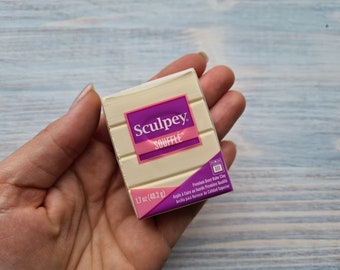 Pode incluir: Uma mão segurando um pacote de argila polimérica Sculpey Soufflé de cor creme. A embalagem tem um rótulo rosa e roxo com o nome da marca e detalhes do produto. Pesa 48,2 gramas.