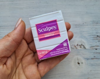 Pode incluir: Uma embalagem de argila polimérica branca Sculpey Soufflé, segurada por uma mão. A embalagem tem um rótulo rosa e roxo com o texto "Sculpey Soufflé" e "1,7 oz (48,2 g)". Também inclui texto em francês e espanhol.