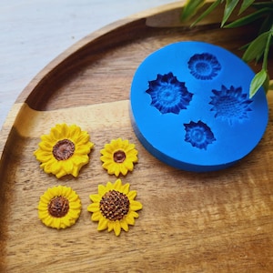 Op de afbeelding: Een blauwe siliconen mal met vijf zonnebloemvormen en vijf gele zonnebloemdecoraties op een houten oppervlak.