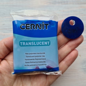 May include: A package of Cernit translucent polymer clay in a blue and white package. A dark blue, circular piece of clay with a hole is next to the package. The package has the word "TRANSLUCENT" in large, green letters.