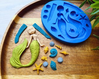 May include: A wooden tray holds a blue silicone mermaid mold, a mermaid figurine, and various sea-themed embellishments. The mermaid figurine has a green tail and a tan torso. Seashells and a starfish are scattered around.