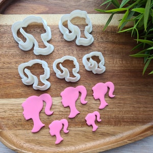 Peut inclure: Un ensemble d'emporte-pièces en plastique blanc en forme de tête de femme avec différentes coiffures. Il y a aussi des découpes en pâte de sucre rose de la même forme.