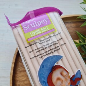 May include: A package of Super Sculpey Living Doll polymer clay. The package is pink and white with the text "super Sculpey Living Doll" on the front. The text on the package reads "Semi-translucent, smooth blending, and perfect for fine detail. Semi-translucide, facile &agrave; m&eacute;langer et id&eacute;al pour cr&eacute;er de petits d&eacute;tails. Semitransl&uacute;cida, mezcla homog&eacute;nea y perfect para los peque&ntilde;os detalles."