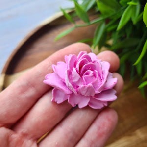 May include: A single pink flower with multiple layers of petals, resembling a rose or peony. The flower is made of a soft, pliable material, possibly polymer clay or resin.