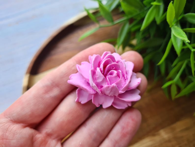 May include: A single pink flower with multiple layers of petals, resembling a rose or peony. The flower is made of a soft, pliable material, possibly polymer clay or resin.