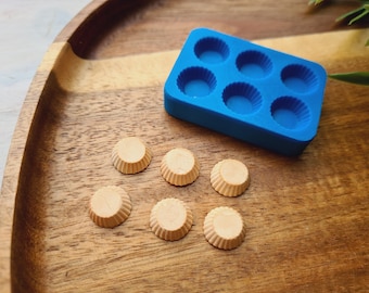 Puede incluir: Un molde de silicona azul con seis cavidades redondas para hacer pequeñas golosinas, junto con seis golosinas redondas terminadas de color marrón claro sobre una superficie de madera.