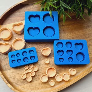 Op de afbeelding: Een houten dienblad toont blauwe siliconen mallen in hart- en cirkelvormen, samen met gebakken taartjes. De mallen zijn voor het knutselen, met verschillende taartjes al gemaakt.