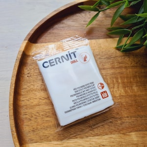 May include: A package of Cernit Doll polymer clay, a skin effect clay, is displayed on a wooden tray. The white package has text in multiple languages, including English, French, and German. Green leaves are in the background.
