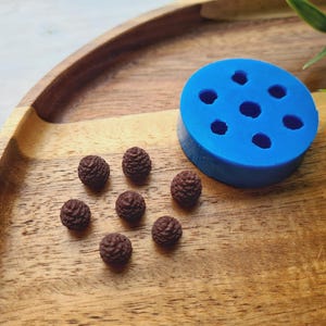 Può includere: Uno stampo in silicone blu con otto cavità rotonde su una superficie in legno. Accanto allo stampo ci sono otto piccole fusioni in resina marrone di pigne.