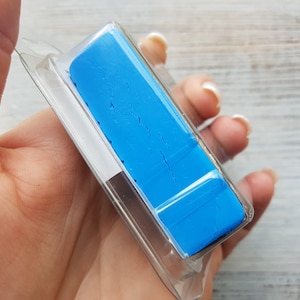 May include: A bright blue art eraser, rectangular in shape, encased in a clear plastic package. The eraser's surface shows cracks. The package is held in a hand, with a light-coloured wooden surface in the background.