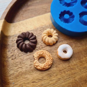 Può includere: Quattro tipi diversi di ciambelle su una superficie di legno. Le ciambelle sono marroni, bianche e beige. C'è uno stampo per ciambelle in silicone blu sullo sfondo.
